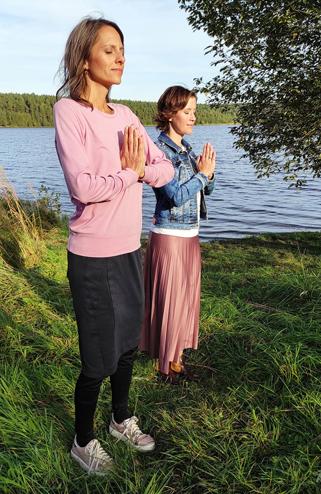 Die Gründerinnen Katrin und Franziska stehen vor einem See. Sie hören eine Fantasiereise und stehen entspannt mit der Meditationspose Händen vor dem Herzen.