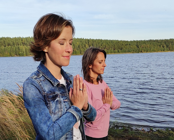 Zwei Frau halten ihre Hände vor dem Herzen und entspannen mit einer Fantasiereisen-Übung. Sie stehen in der Natur vor einem See.